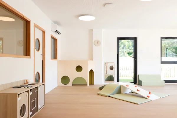 Salle de jeux de rôles de la crèche Les Bébés Explorateurs Limonest (69670) – cuisine dinette bois et marron, maison tunnel arches vertes avec hublots, rampe motricité vert sauge, miroir sur pied et banquette vert menthe