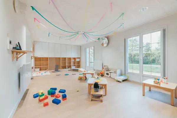 Salle de motricité de la crèche Les Bébés Explorateurs Vertou Matisse (44120) – grandes briques Lego colorées, rubans colorés suspendus au plafond, étagères bois blanches et grandes baies vitrées sur jardin