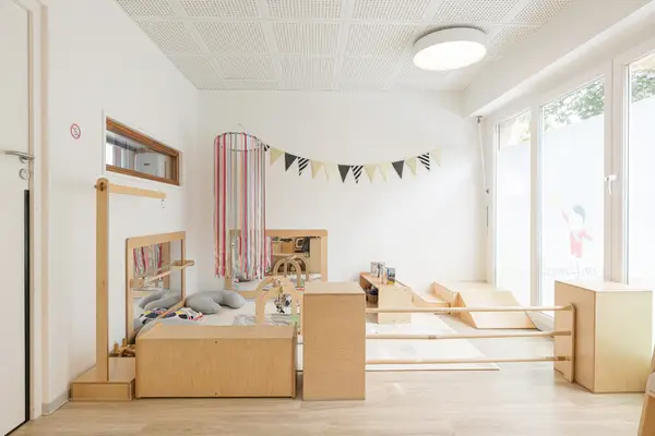 Salle bébés de la micro-crèche Les Bébés Explorateurs Orléans Saint-Marceau (45100) – bac à jouets bois, portique à barres colorées, plan incliné bois, guirlande fanions noir et blanc, grande fenêtre sur verdure